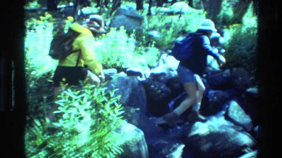 Two backpacked hikers, one in a yellow jacket and one in a blue shirt, move over boulders in a bright wooded stream setting.