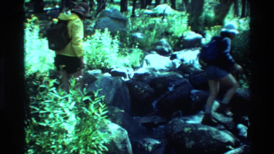 Backpacked hikers continue across uneven boulders in a wooded British Columbia setting, with bright foliage and soft 8mm color shifts.