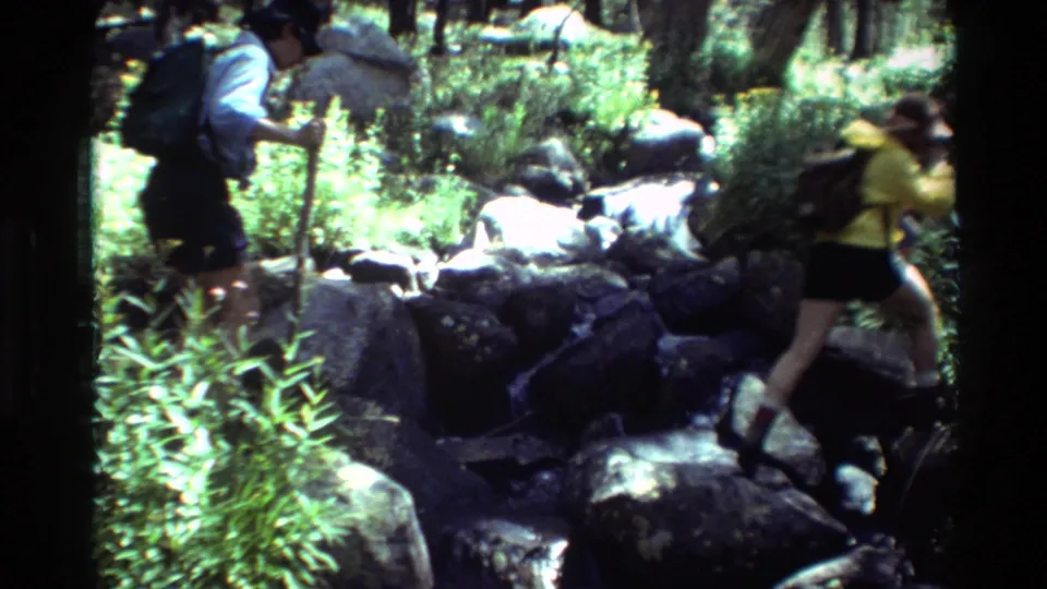 1981 8mm frame of two backpacked hikers navigating rocks and vegetation in a forest stream crossing.
