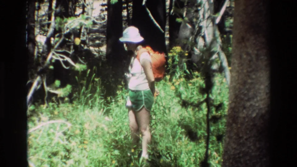 A hiker wearing a light hat and red backpack walks away through a grassy forest area in metadata-described British Columbia footage.