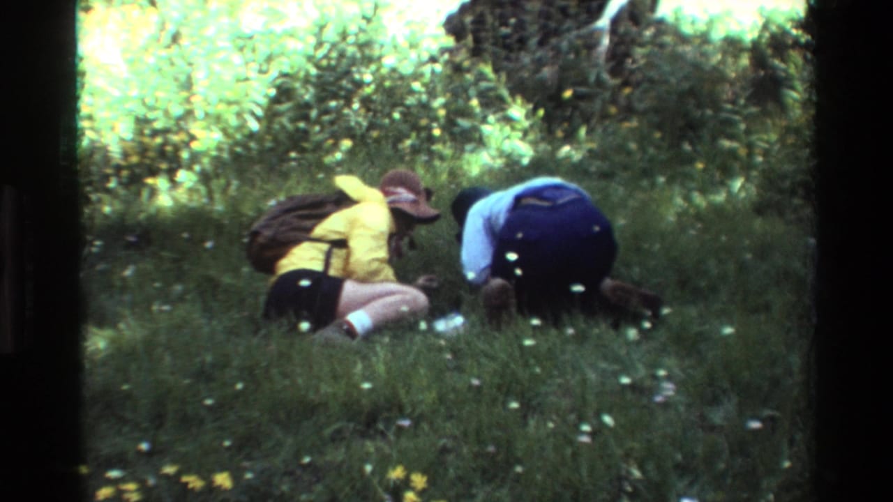 1981 8mm frame from supplied British Columbia metadata showing two adults in hats crouched in tall grass during a field observation.