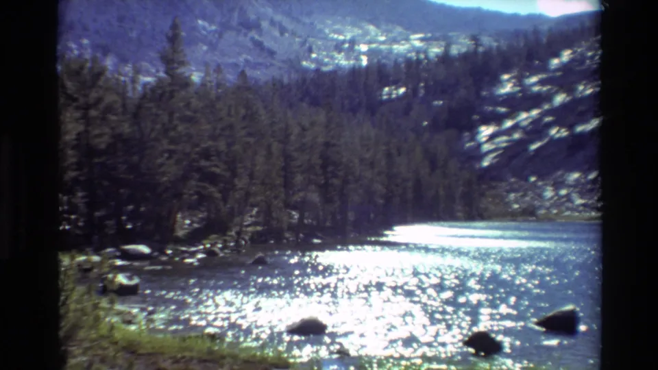 Metadata-supported British Columbia 8mm frame of a river bordered by trees and rocks, with strong sun glare on the water.