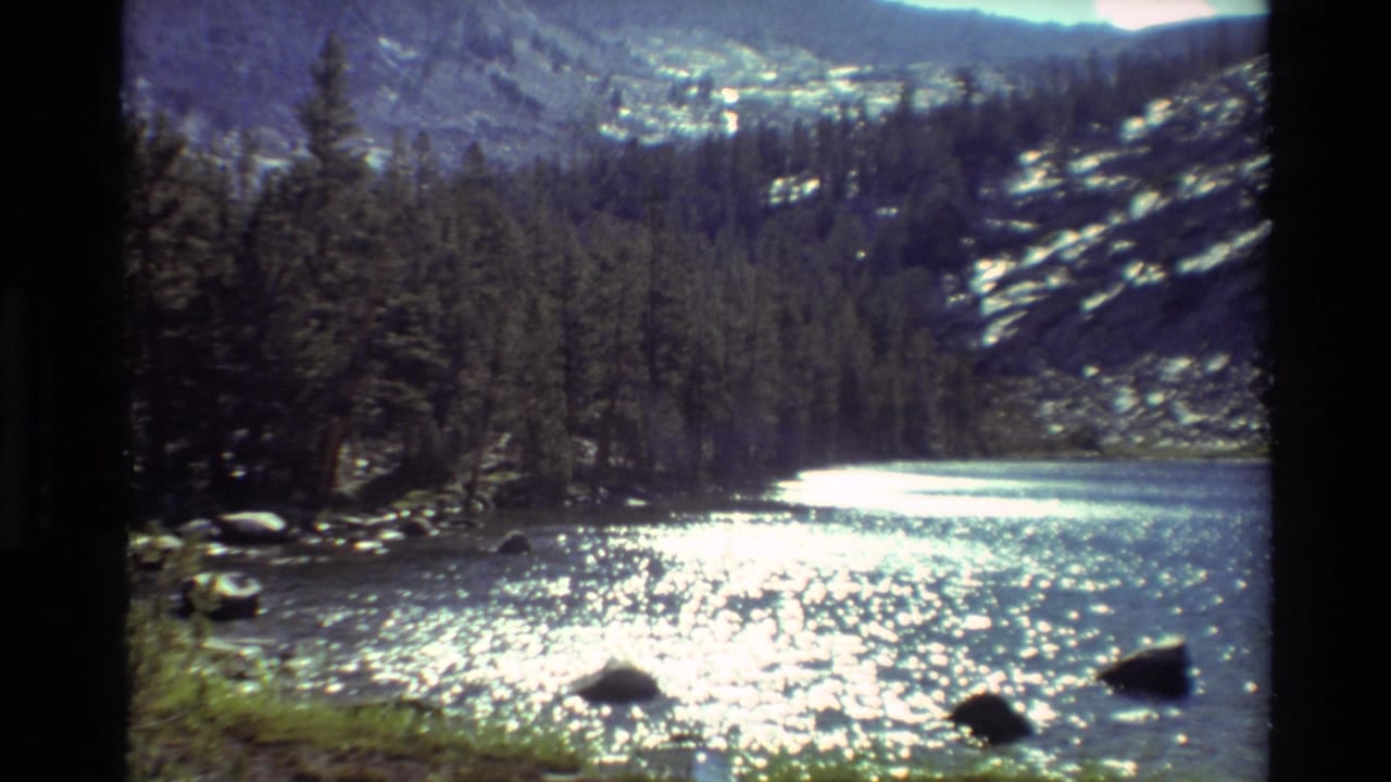 Sunlit lake with sparkling water, dark shoreline rocks, evergreen trees, and rocky slopes beyond.