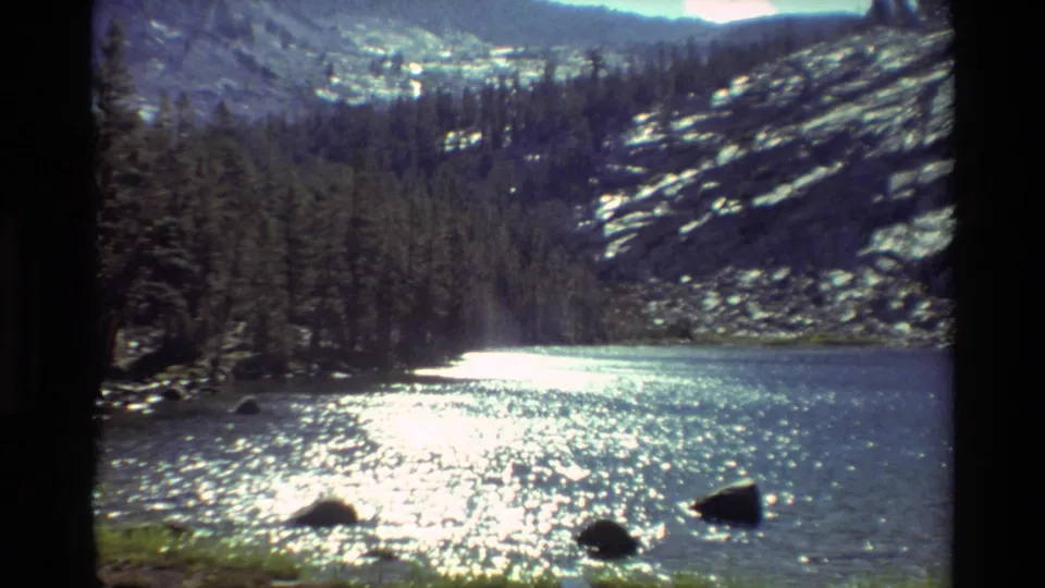 1981 8mm lakeside frame with intense sun reflection on water, dark rocks in the foreground, evergreen trees, and pale rocky mountain slopes.