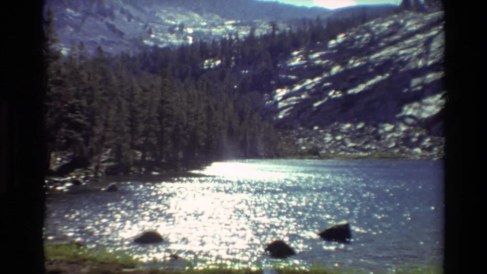 Final 1981 8mm frame of a quiet lake scene, with sparkling water, shoreline rocks, conifers, and rugged mountain terrain beyond.