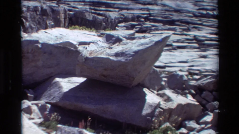1981 8mm frame showing flat layered rocks, small pebbles, and a shallow water edge in a natural outdoor setting.