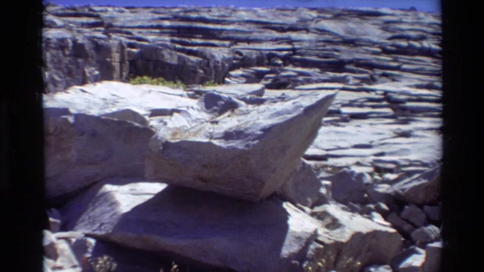 1981 8mm frame of a rocky shoreline with broad flat stones, pebbles, and layered rock in bright daylight.
