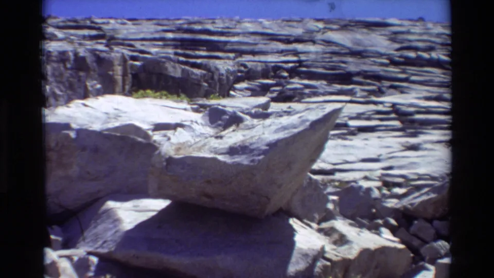 1981 8mm frame of stratified rock and an angular boulder in a sunlit metadata-labeled British Columbia/USA landscape.