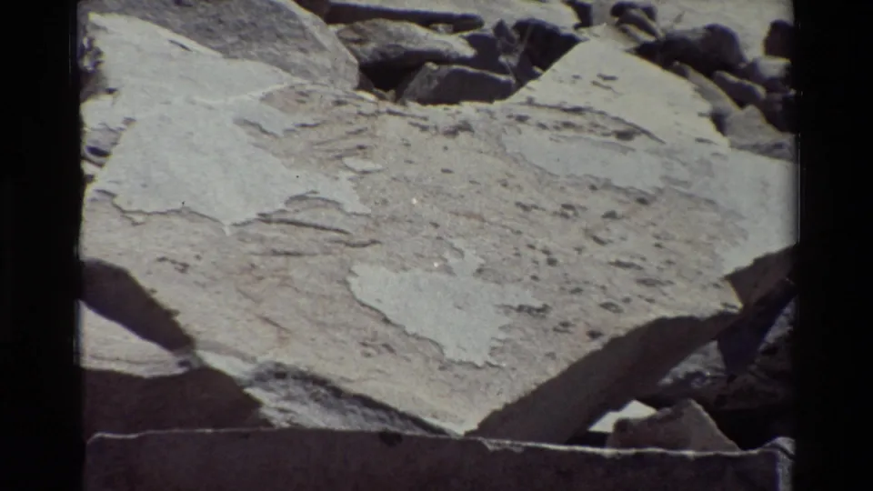 Close 8mm view of flat pale stone, rough surface markings, and darker rocks at the top edge of the frame.