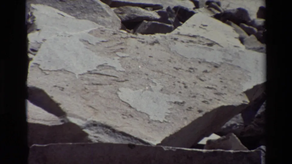 Handheld 8mm frame of pale layered rock, broken edges, and scattered darker stones in an outdoor rocky area.