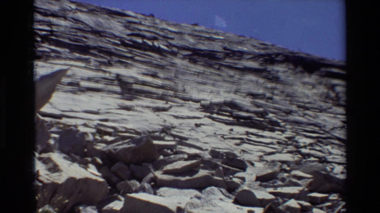 1981 8mm frame cataloged to British Columbia showing a stratified rock wall, rubble slope, and pale boulders in hard sunlight.