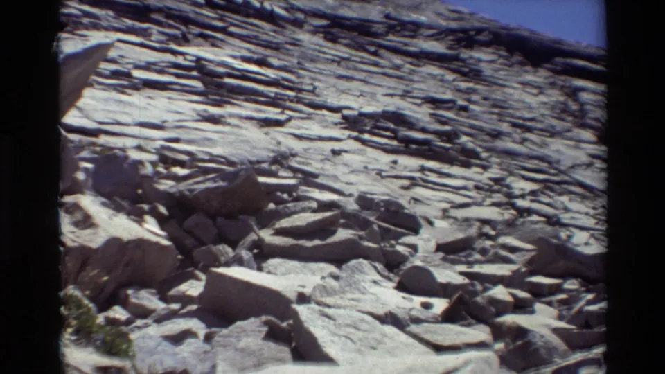 Cataloged 1981 outdoor home-movie frame with a bright stratified rock face, broken boulders, and a small patch of vegetation near the lower edge.