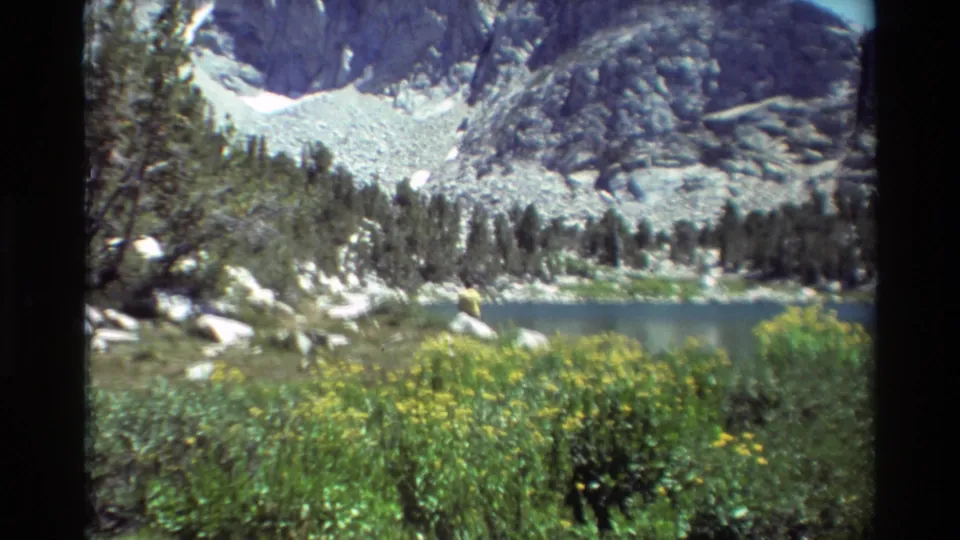 1981 8mm frame of a blue alpine lake below rocky slopes and snow patches, with evergreens and wildflowers in the foreground.