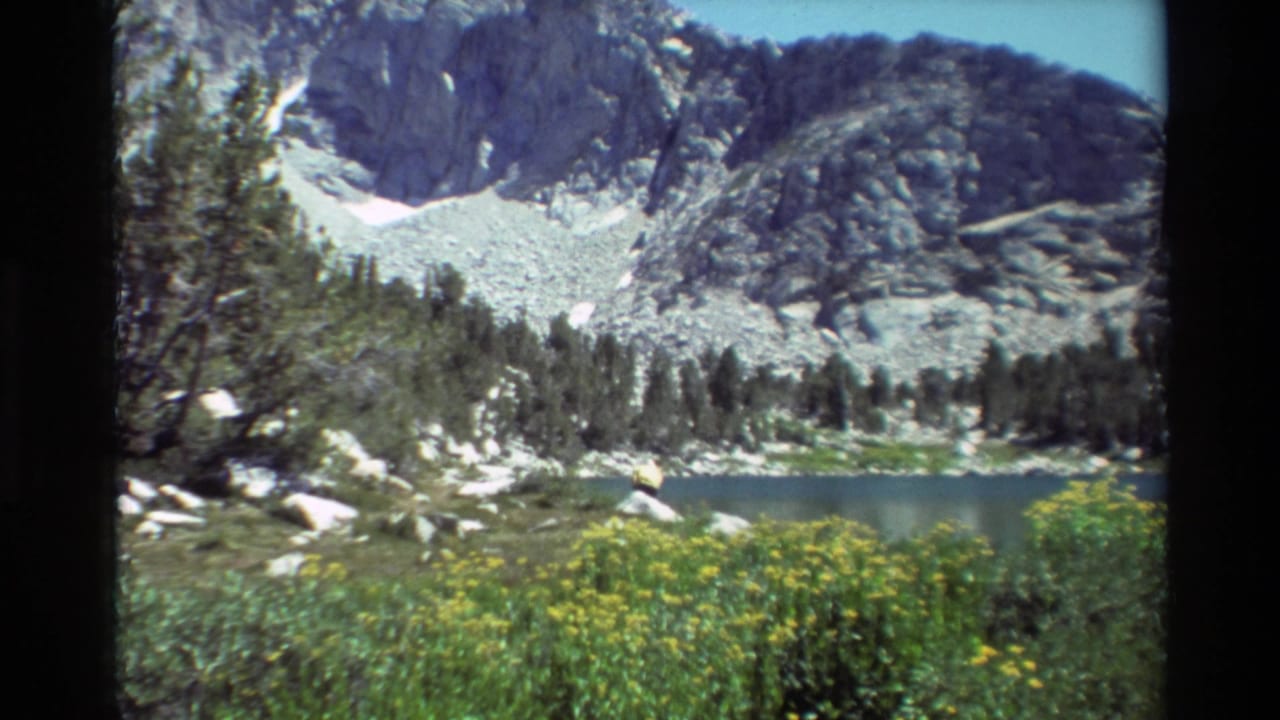 1981 8mm frame of an alpine lake scene, with evergreen trees, yellow wildflowers, boulders, and a snow-streaked rocky mountain behind.