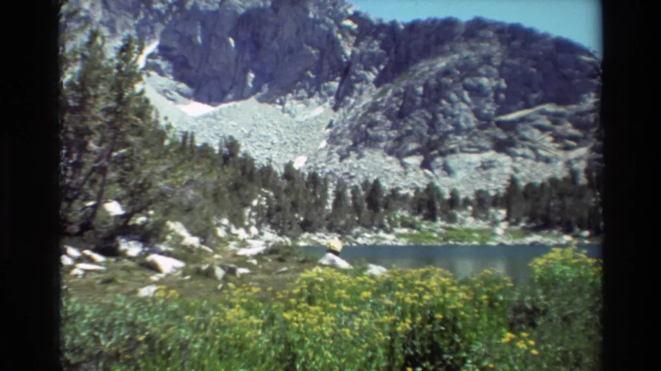 1981 8mm frame of an alpine lake scene, with evergreen trees, yellow wildflowers, boulders, and a snow-streaked rocky mountain behind.
