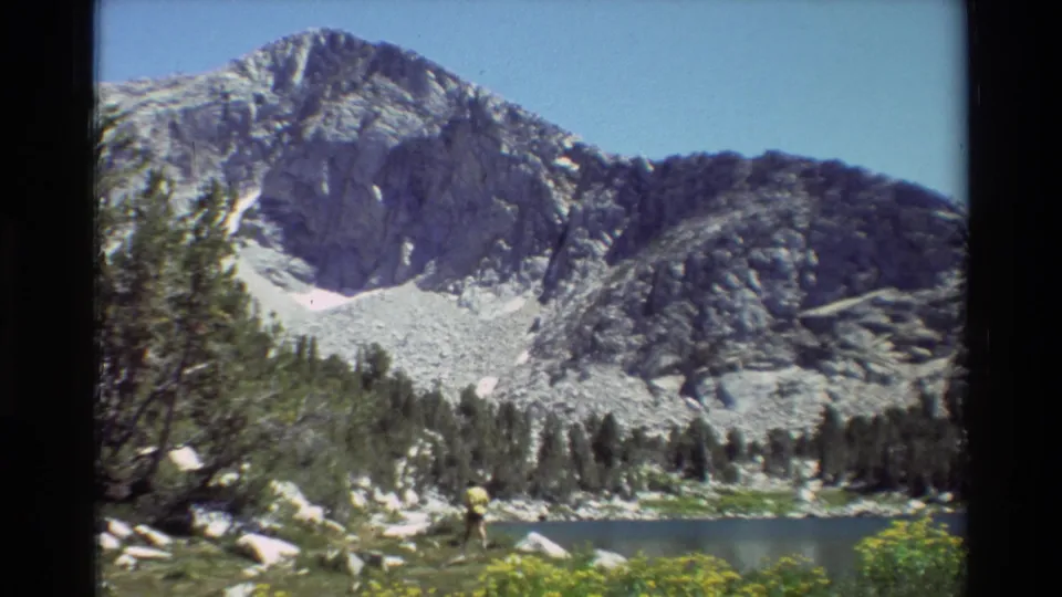 The frame shows a distant hiker beside an alpine lake, backed by evergreen trees, boulders, blue sky, and a snow-marked mountain slope.