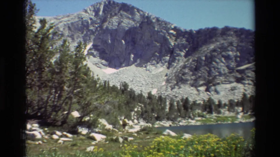 A hiker in yellow appears near the lakeshore in archival 8mm footage, with yellow wildflowers, evergreens, boulders, and a rugged mountain slope behind.