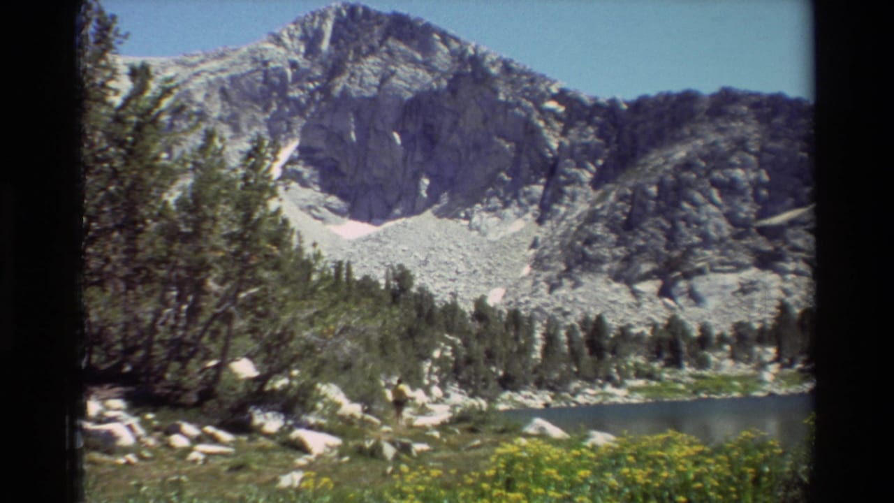 1981 8mm frame of a rocky alpine peak rising above a small lake, with evergreens, boulders, and yellow wildflowers in the foreground.