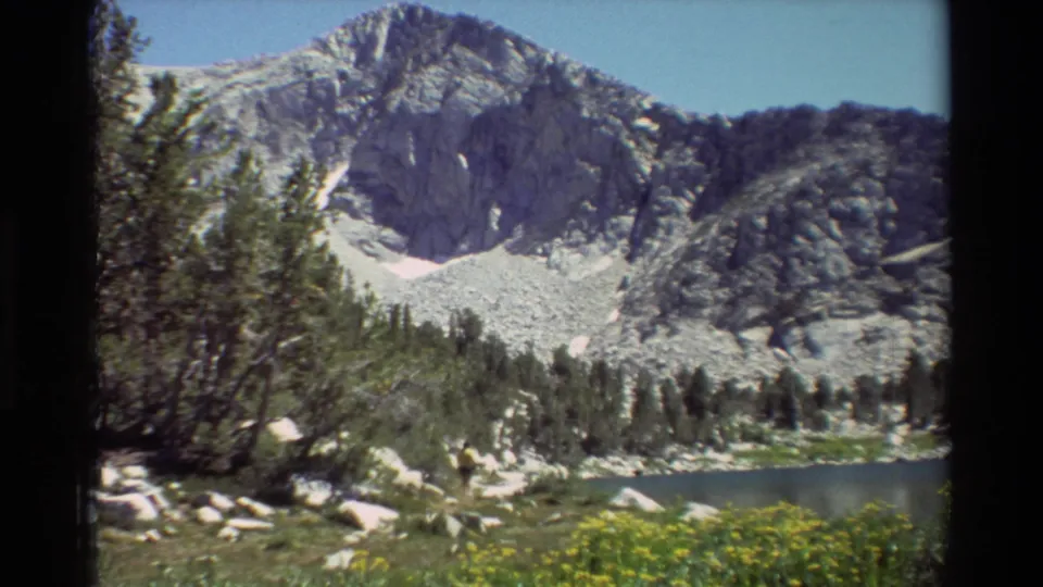 1981 8mm frame of a rocky alpine peak rising above a small lake, with evergreens, boulders, and yellow wildflowers in the foreground.