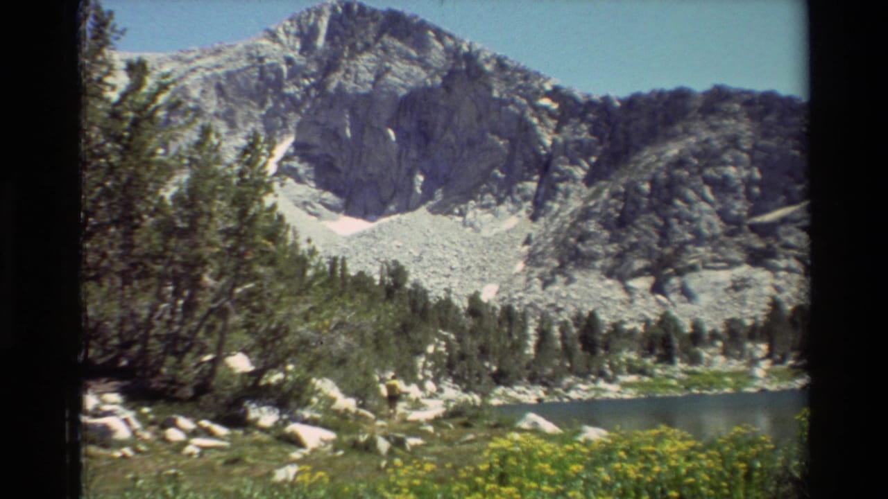 Vintage 8mm view of a rugged mountain basin with a calm lake, conifers, pale rocks, and meadow growth in the foreground.