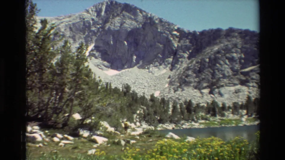 Vintage 8mm view of a rugged mountain basin with a calm lake, conifers, pale rocks, and meadow growth in the foreground.