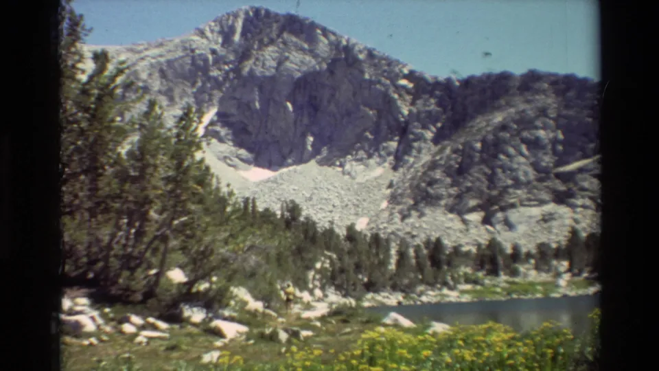 1981 8mm mountain landscape showing a steep rocky peak, conifer line, small lake, scattered boulders, and yellow wildflowers.
