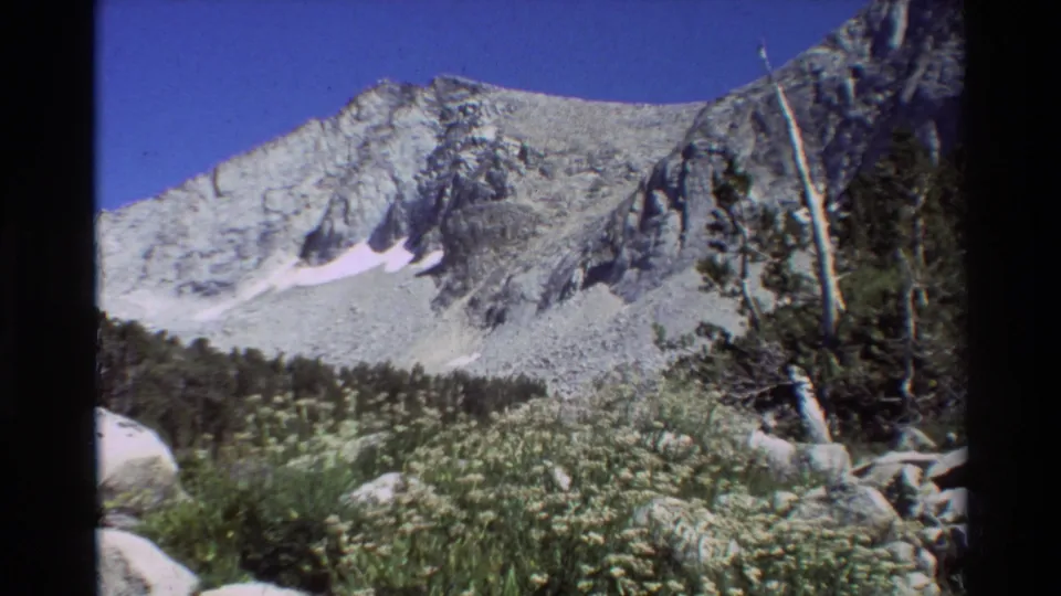 Wide home-movie frame of rocky mountain peaks, sparse trees, pale rocks, and alpine vegetation in the listed British Columbia location.