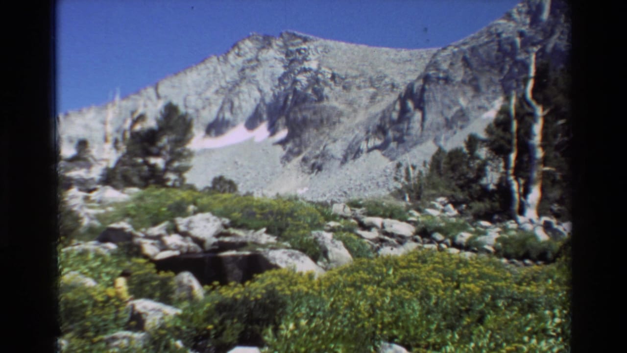 1981 8mm home-movie view of a rugged alpine basin with pine trees, boulders, shrubs, snow patches, and sunlit rock walls.