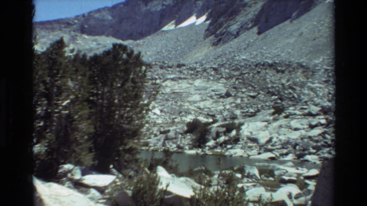 Small mountain lake below rocky slopes with trees, boulders, snow patches, and blue sky.