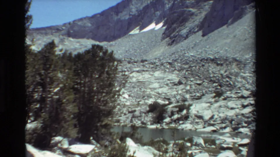 1981 8mm home-movie view across trees and rocks toward a small mountain lake, talus slopes, and bright snow patches.