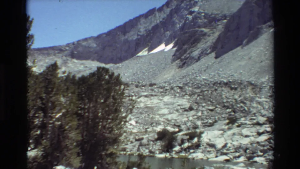 1981 8mm home-movie frame showing a small lake below conifers, pale boulders, rocky slopes, and late-season snow patches.