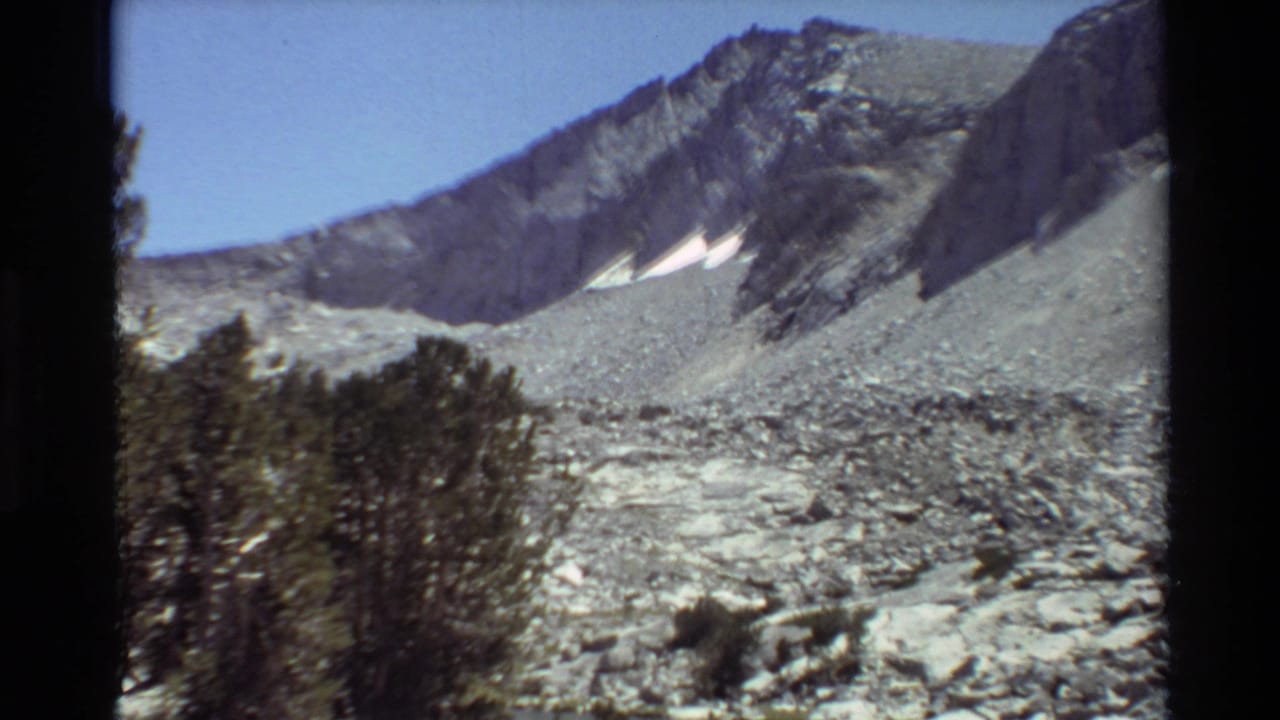 Cataloged 1981 8mm frame of a rugged mountain slope with small snow patches, evergreen foreground, blue sky, and visible film borders.