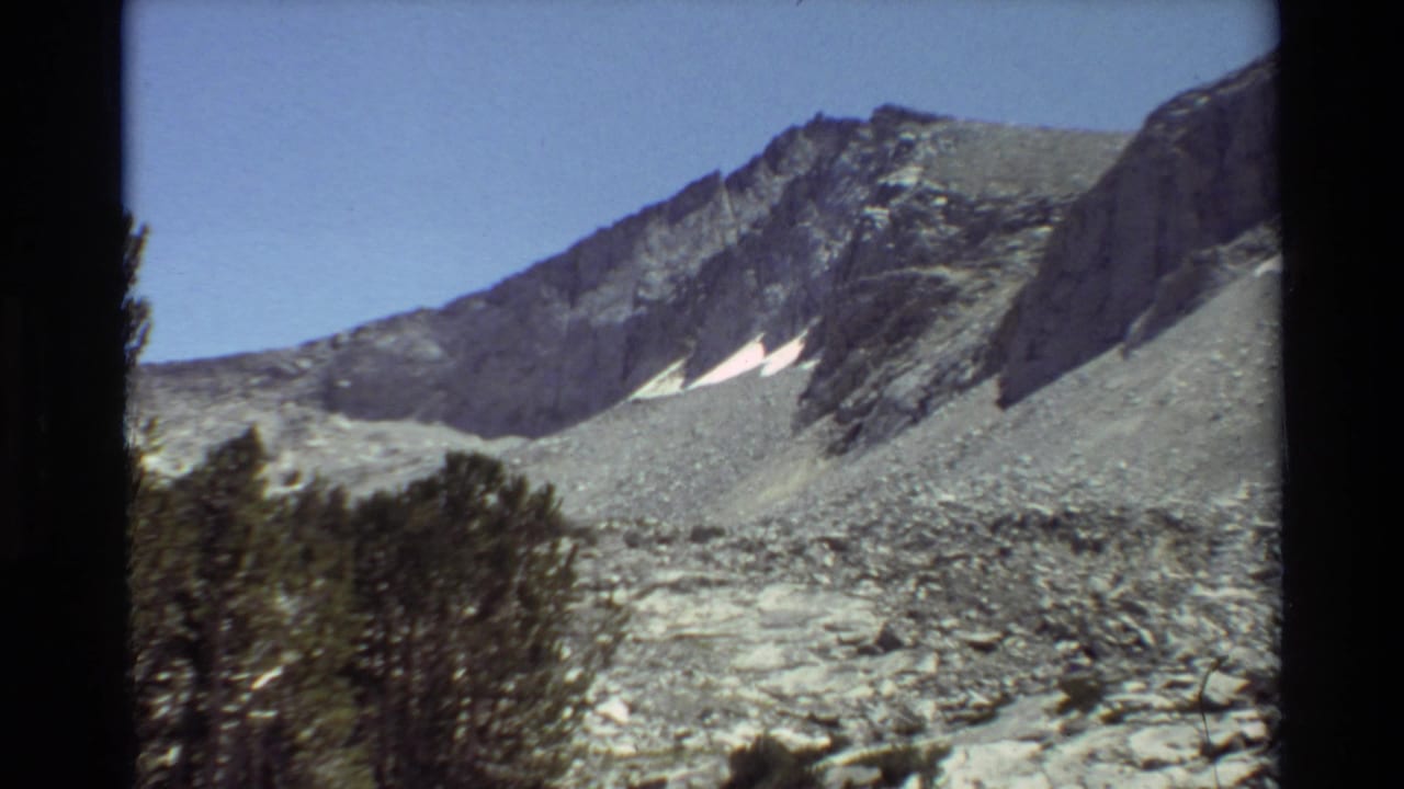 Cataloged 1981 8mm view of jagged gray peaks, a pale rocky slope, small snow patches, and evergreen trees near the frame edge.