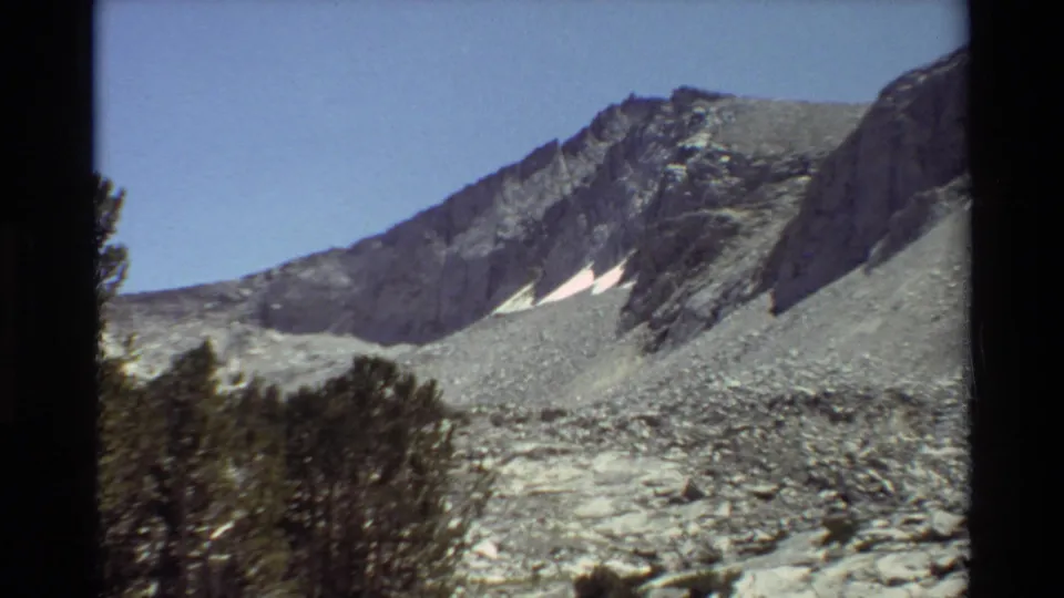 Cataloged 1981 home-movie frame showing a rocky alpine ridge, lingering snow, evergreen foreground, and visible 8mm gate edges.