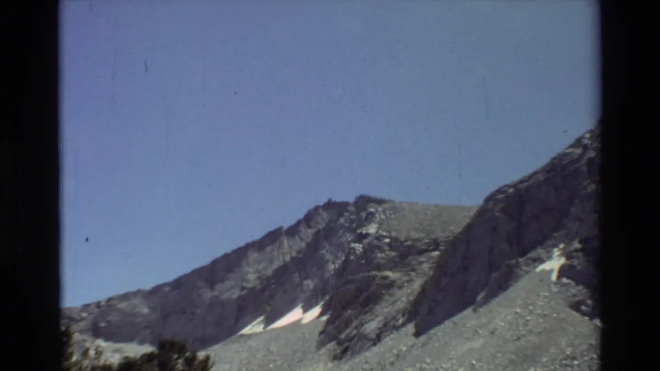 Cataloged 1981 8mm frame with a wider view of rocky alpine slopes, blue sky, small snow patches, and dark scan borders.
