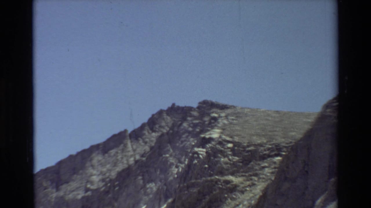1981 8mm home-movie frame moving closer to a jagged rocky summit area, with textured stone against blue sky.