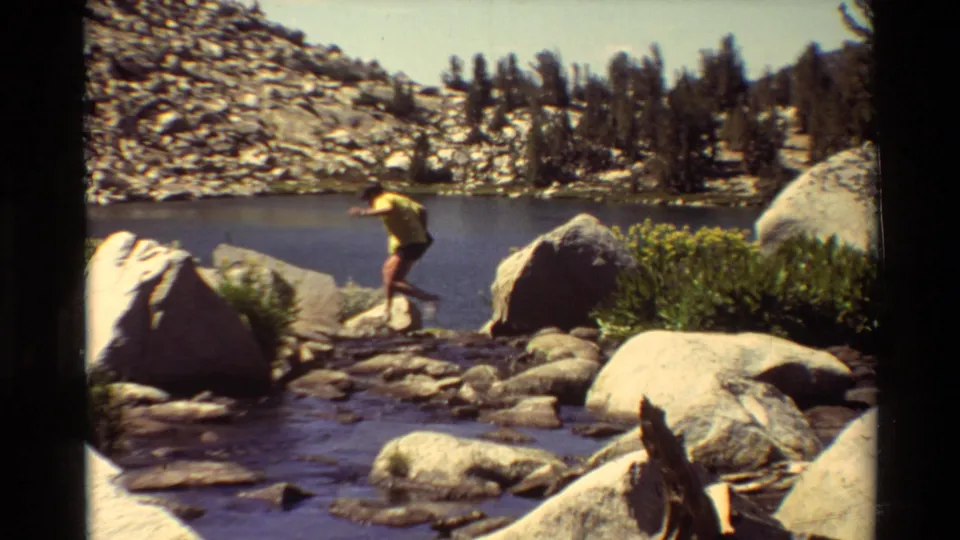 A man in a yellow shirt and dark shorts crosses shoreline rocks beside a mountain lake in 1981 home-movie footage.