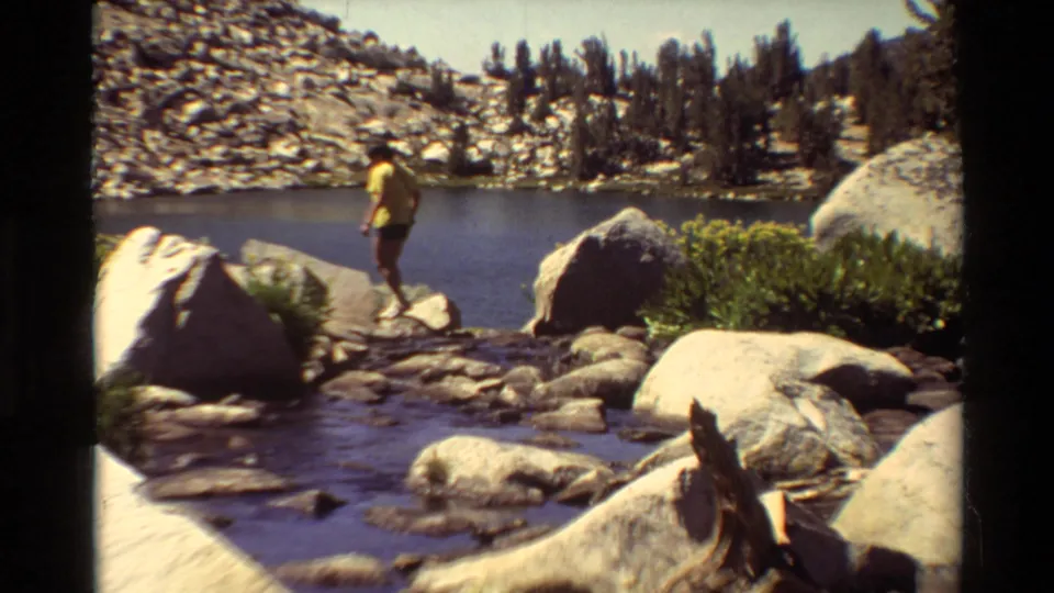 The man continues across the rocky lake edge, framed by boulders, water, evergreens and rugged slopes in a 1981 8mm scene.
