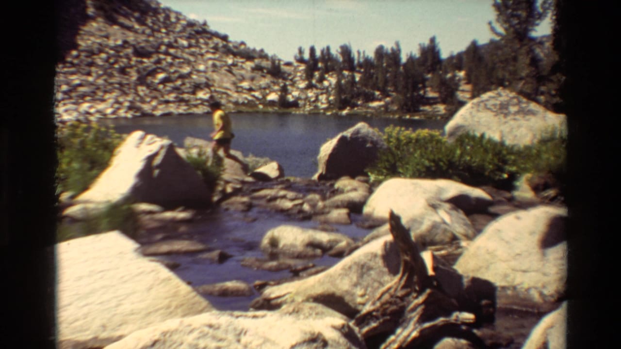 1981 8mm frame of a person in yellow crossing rocks near a shallow mountain stream, with a small lake and rocky slope behind.
