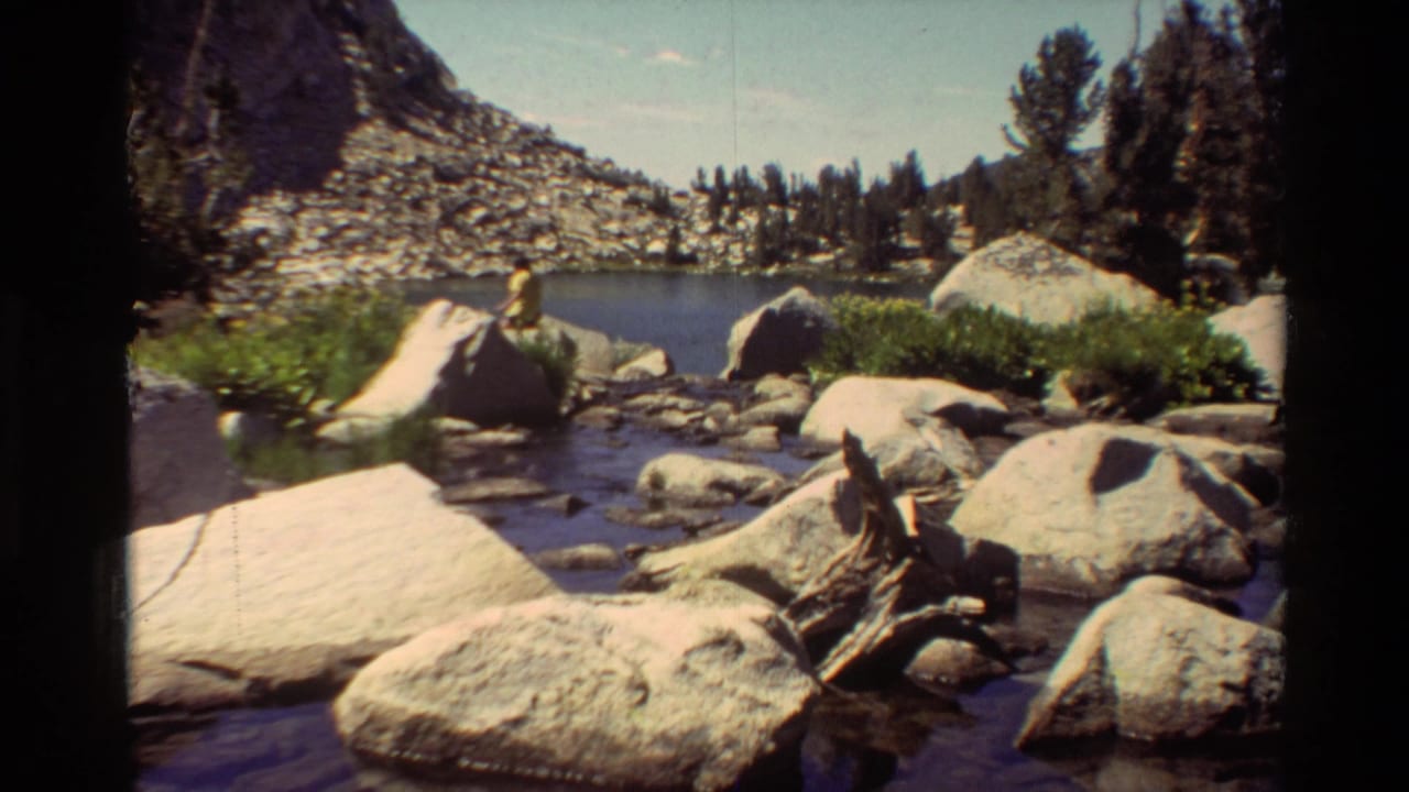 Person partly visible near boulders by a shallow stream, with lake and trees in the background.