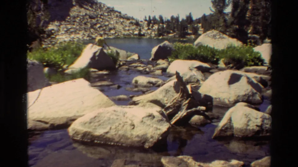 8mm frame of a shallow rocky stream leading toward a mountain lake, with boulders, green shrubs, pines, and a rocky slope.