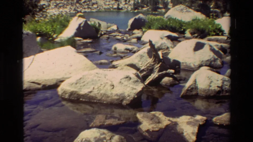 Sunlit 8mm view of a rocky mountain stream with driftwood, large boulders, lakeshore greenery, pines, and a stony hillside.