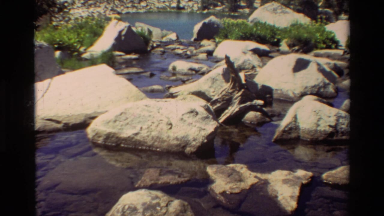1981 8mm home-movie frame of a rocky mountain stream with clear water, sunlit boulders, and a partially submerged log.