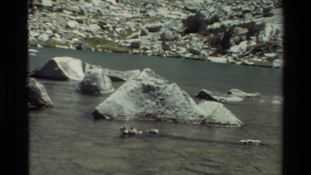 1981 8mm frame showing a calm rocky river channel with pale boulders, a stony far bank, and sparse shrubs.