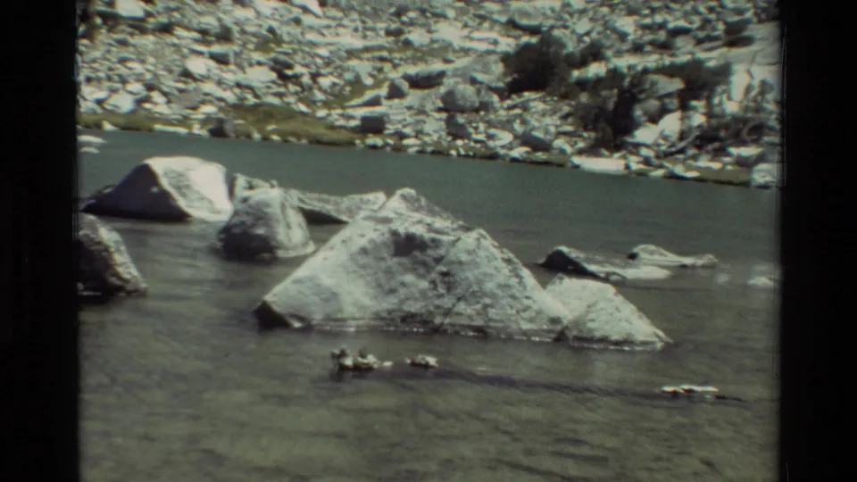 1981 8mm frame showing a calm rocky river channel with pale boulders, a stony far bank, and sparse shrubs.