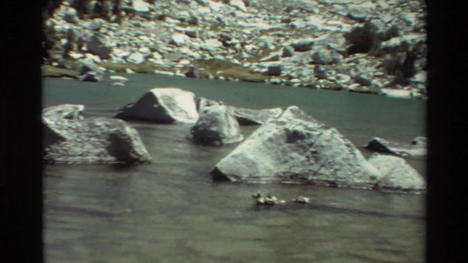 1981 8mm home-movie view of a shallow mountain river passing around pale boulders below a rocky bank.