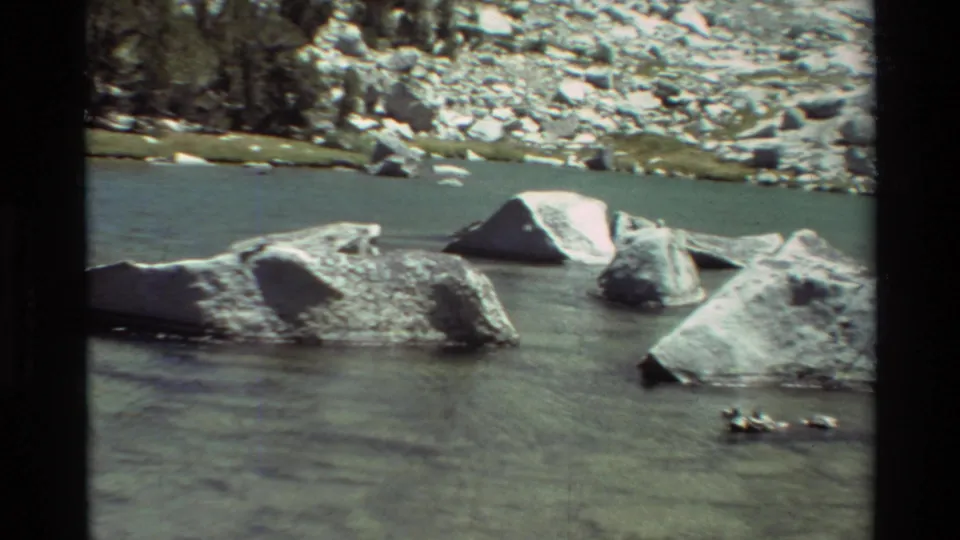 1981 8mm frame of flowing river water, rounded boulders, and a rocky mountain-bank backdrop.