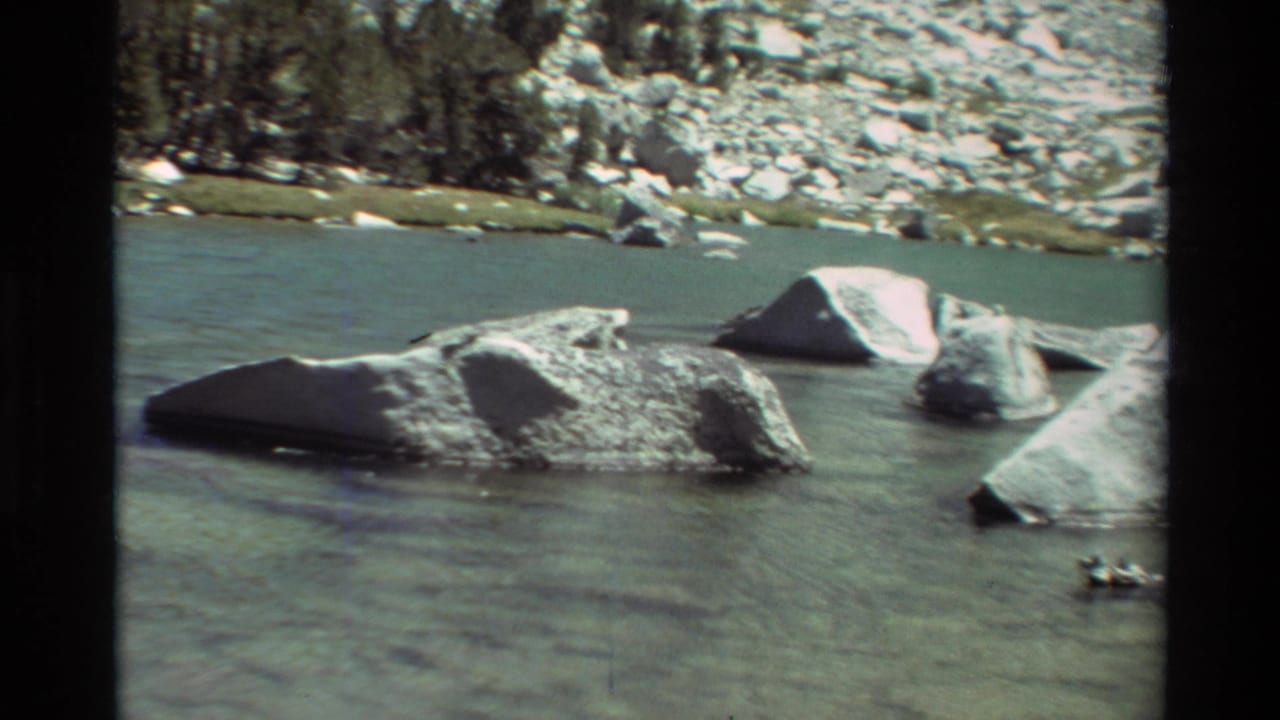 1981 8mm frame of partly submerged boulders in a calm mountain lake, with rocky shore and evergreens beyond.