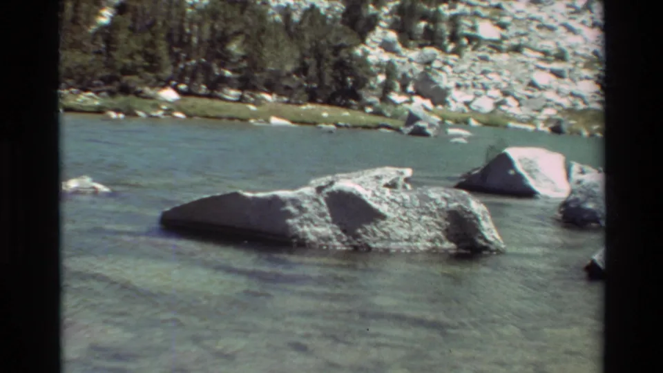 Mountain lake view from supplied British Columbia metadata, showing shallow water, large shoreline rocks, and evergreen trees on 8mm film.