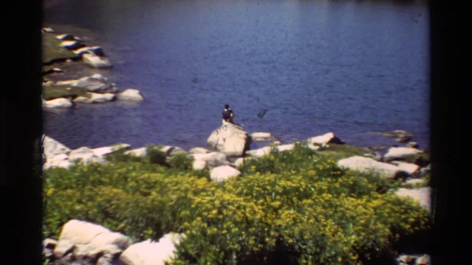 Home-movie frame of a distant lakeside figure on a boulder, with blue water, pale rocks, and yellow wildflowers under sun.